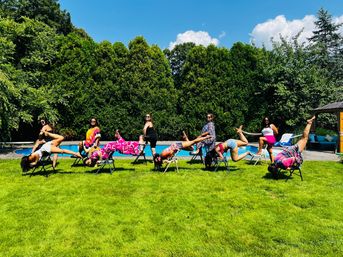 Playful group of women doing upside-down and balancing chair poses on folding chairs on a sunny backyard lawn beside a swimming pool with tall hedges and blue sky