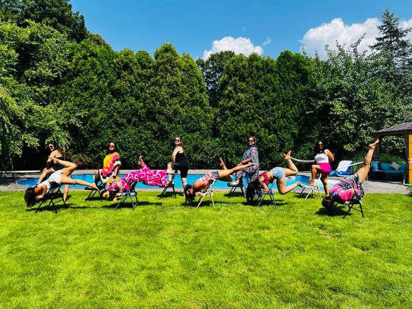 Playful group of women doing upside-down and balancing chair poses on folding chairs on a sunny backyard lawn beside a swimming pool with tall hedges and blue sky