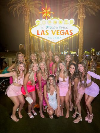 Smiling group of friends in pink outfits posing at night in front of the illuminated Welcome to Fabulous Las Vegas Nevada sign, framed by palm trees and Strip lights