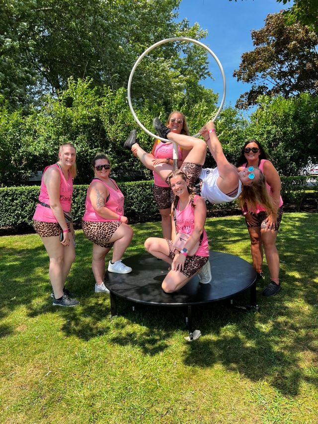 Six women in matching pink tank tops and leopard-print shorts pose around a white aerial hoop on a circular platform in a sunny park with green trees and blue sky, one woman hanging upside down from the hoop.