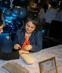 Smiling vendor in a denim jacket fastening a delicate gold bracelet on a customer's wrist at a lively indoor pop-up jewelry table with blue balloon decorations and a framed price sign.