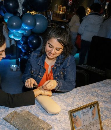 Smiling vendor in a denim jacket fastening a delicate gold bracelet on a customer's wrist at a lively indoor pop-up jewelry table with blue balloon decorations and a framed price sign.