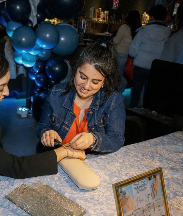 Smiling vendor in a denim jacket fastening a delicate gold bracelet on a customer's wrist at a lively indoor pop-up jewelry table with blue balloon decorations and a framed price sign.