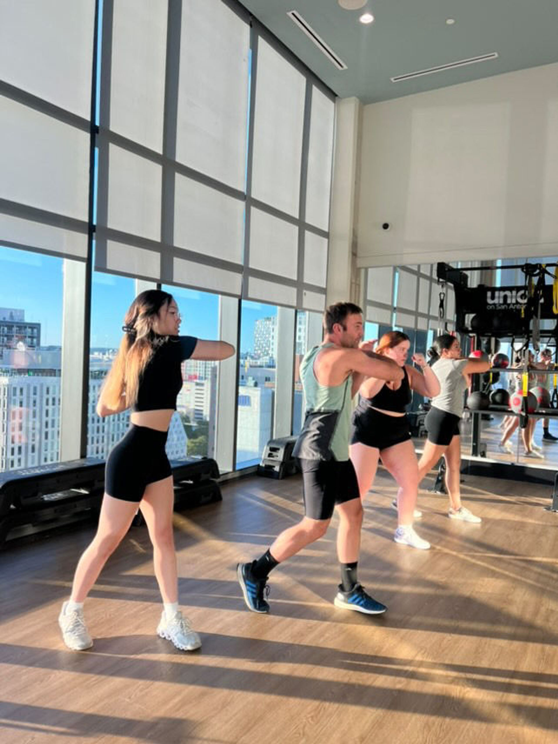 Four people in a bright high-rise gym studio with floor-to-ceiling windows and city skyline, doing a group fitness boxing-style workout and throwing synchronized punches in front of mirrors.