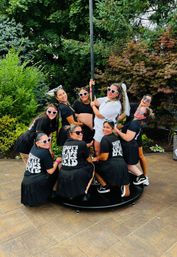 Lively outdoor bachelorette party on a backyard patio with a bride in white and veil surrounded by friends in matching black shirts and heart-shaped sunglasses posing around a pole, lush garden trees in the background.