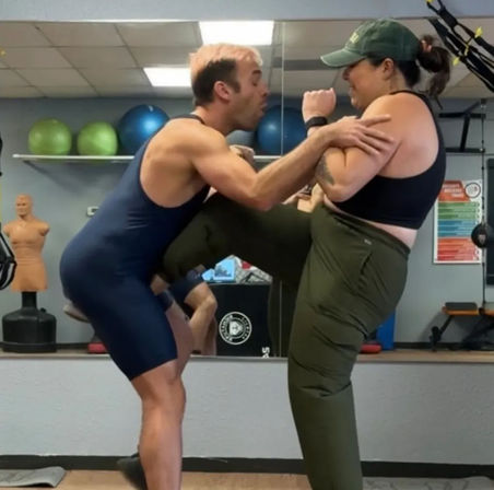 Two adults in a fitness studio practicing self-defense: woman in green pants delivers a knee strike to a trainer in a navy singlet in front of a mirror, with stability balls and a training dummy in the background.