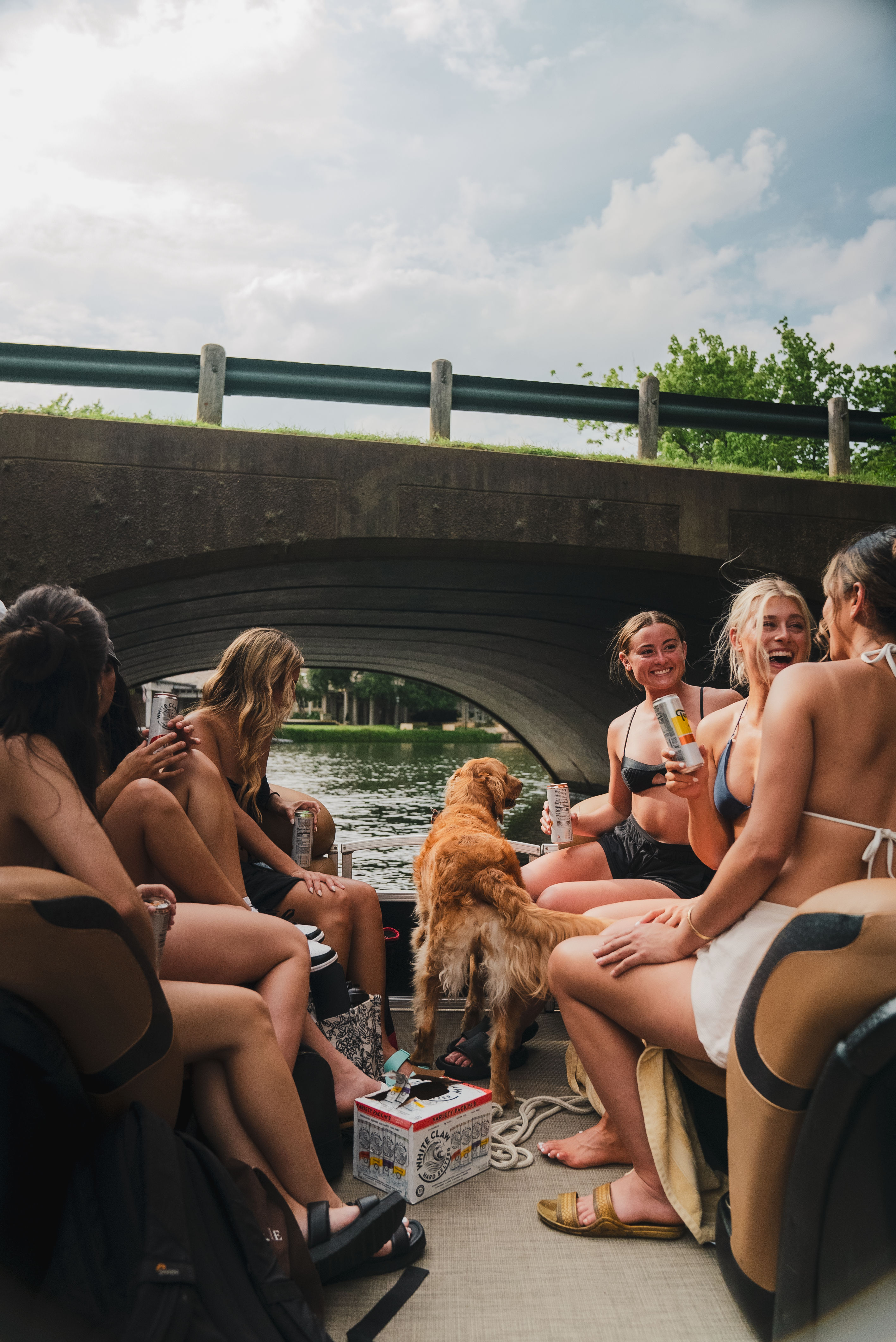 Young friends in swimsuits on a pontoon boat under a concrete bridge on a summer river, laughing and sipping canned drinks with a golden retriever aboard.