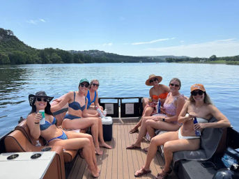 Six friends in swimsuits relaxing with drinks on a pontoon boat on a sunny lake with tree-lined shores and blue sky — summertime boating outing.