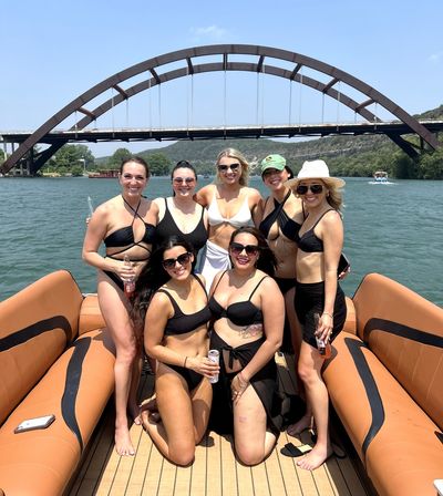 Group of seven friends in swimsuits smiling on a pontoon boat under a large arched steel bridge over a sunny lake with wooded hills in the background