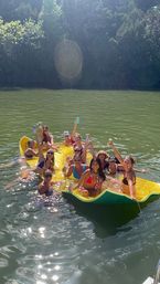 Group of friends relaxing on a large yellow inflatable raft at a tree-lined lake, smiling and raising drinks on a sunny summer day.
