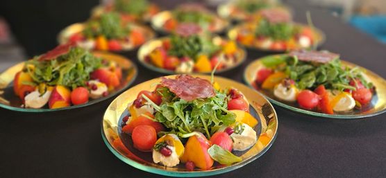 Vibrant plated summer salads on gold-rim plates: arugula piled with a slice of cured meat, watermelon balls, peach wedges, creamy cheese dollops and pomegranate seeds on a black tablecloth.