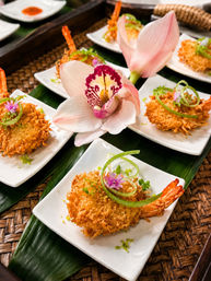 Crispy coconut-crusted shrimp appetizers on small white plates, garnished with green onion curls and purple edible flowers, arranged on a banana leaf with a pink orchid
