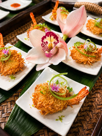 Crispy coconut-crusted shrimp appetizers on small white plates, garnished with green onion curls and purple edible flowers, arranged on a banana leaf with a pink orchid