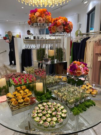 Chic boutique event spread with mirrored tables, vibrant orange and pink rose centerpieces, glowing candles and assorted hors d'oeuvres (mini sliders, cucumber bites, salad cups) beside clothing racks under a crystal chandelier