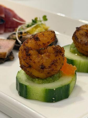 Close-up of a spicy grilled shrimp canapé on a cucumber round topped with avocado mash and bright orange salmon roe, plated as a gourmet seafood appetizer.