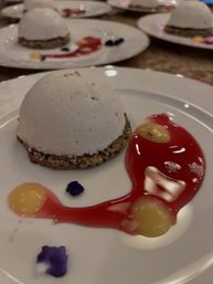 White dome dessert on a crunchy sesame-crumb base, plated with red berry sauce, yellow custard dots and purple edible petals on a white plate