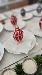 Red-and-gold jeweled Fabergé-style egg on a white plate at an elegant table setting, blurred matching eggs in the background and dipping sauces with greenery in the foreground.