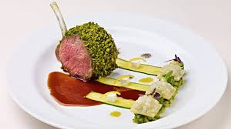 Gourmet herb-crusted lamb chop with a pink medium-rare center, plated with red wine reduction and bright green pea purée topped with diced vegetable terrine on a white plate.
