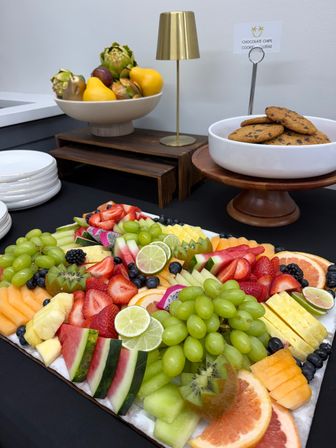 Vibrant catered fruit platter on a black buffet table — watermelon, green grapes, strawberries, pineapple, kiwi and lime slices, with a bowl of chocolate chip cookies and decorative fruit bowl in the background.