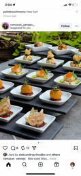 Rows of bite-sized gourmet hors d'oeuvres — seared scallops with roe, fried croquettes, dumplings and wrapped parcels — served in white square dishes on black slate risers in a leafy outdoor event setting, elegant fine-dining appetizers for catered gatherings.