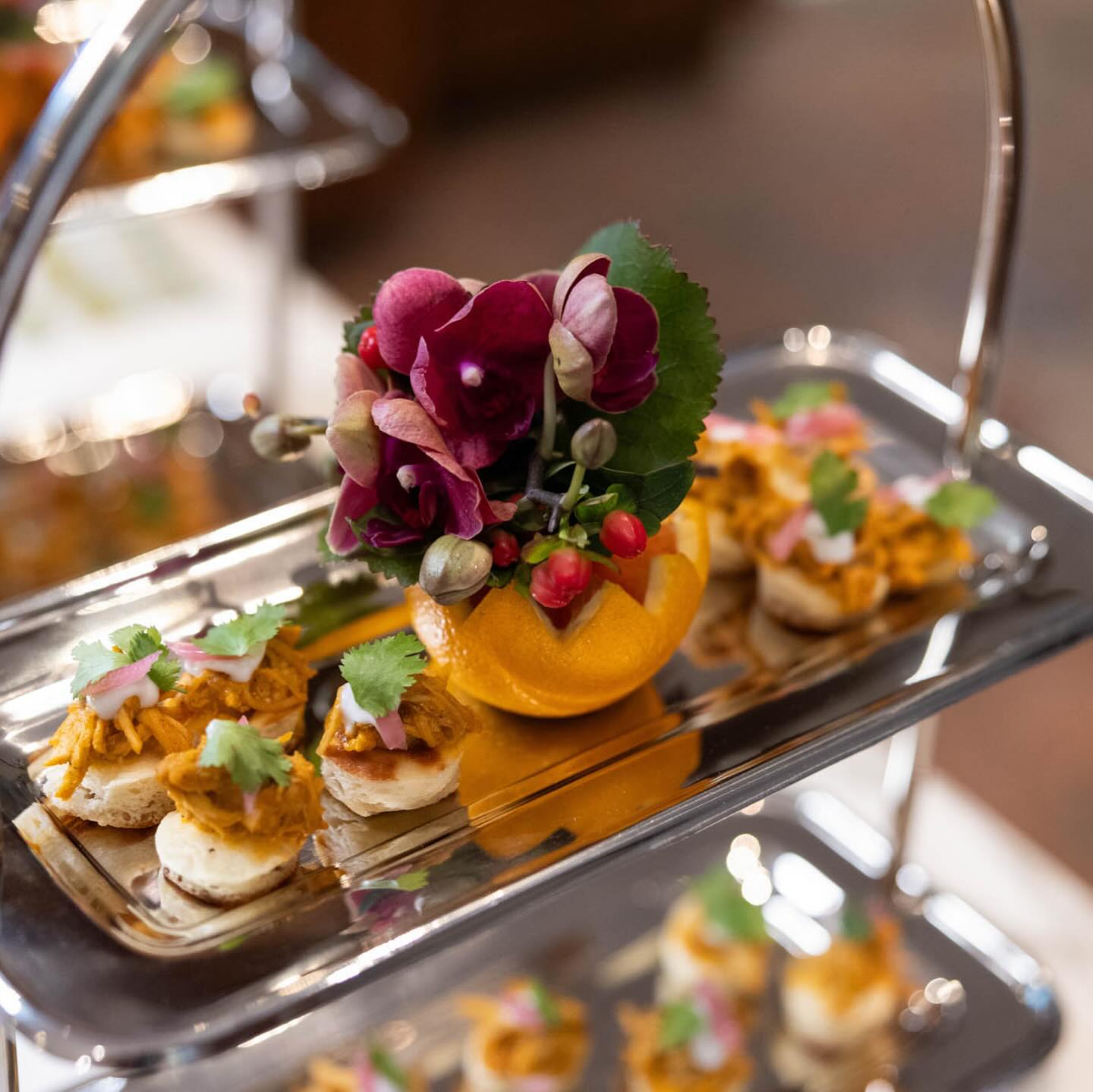 Silver tiered tray of gourmet bite-sized canapés topped with shredded spiced chicken, cilantro and pickled onions, centered around a halved orange filled with purple orchids and berries