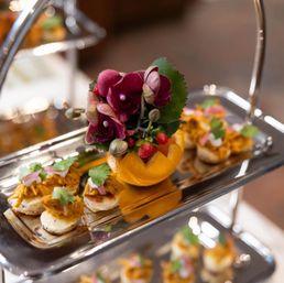 Silver tiered tray of gourmet bite-sized canapés topped with shredded spiced chicken, cilantro and pickled onions, centered around a halved orange filled with purple orchids and berries