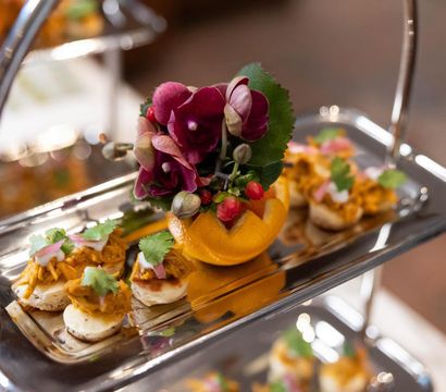 Silver tiered tray of gourmet bite-sized canapés topped with shredded spiced chicken, cilantro and pickled onions, centered around a halved orange filled with purple orchids and berries