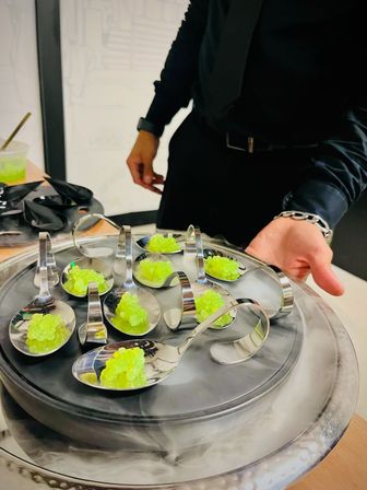 Circular metal tray of silver tasting spoons filled with neon green molecular-gastronomy caviar pearls resting on dry ice, served by a server in black attire — modern gourmet appetizer.