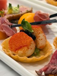 Seared scallop tartlet dusted with paprika, topped with a basil leaf and glossy black pearls in a crisp pastry cup on a white appetizer platter with prosciutto and melon