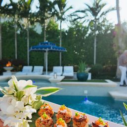 Bite-sized seafood appetizers topped with cream and roe on a platter with white orchids, served poolside at a tropical backyard with a blue umbrella, lounge chairs and palm trees.