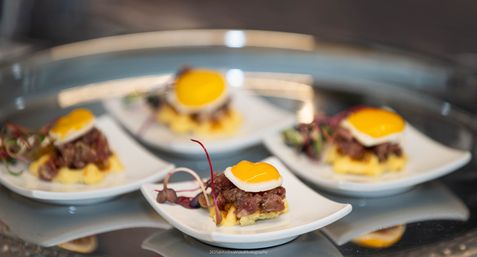 Close-up of bite-sized gourmet canapés: beef tartare on mini polenta squares topped with glossy quail egg yolks and microgreens, plated on white tasting dishes on a mirrored tray.