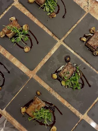 Overhead view of gourmet seared meat squares on black slate tiles, drizzled with dark glaze and garnished with microgreens and roasted pearl onions on a stone countertop.