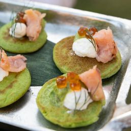 Green blinis topped with smoked salmon, creamy dollops, orange salmon roe and dill on a silver serving tray — elegant smoked salmon canapés for events and catering.