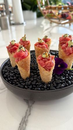 Tuna tartare mini cones topped with wasabi crema and sesame seeds, arranged upright in a bowl of black beans and garnished with a purple edible flower on a marble countertop — gourmet appetizer.