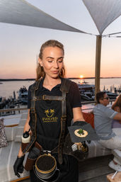 Server in a black apron presenting a caviar-topped canapé on a cracker at a sunset waterfront event by a marina with boats