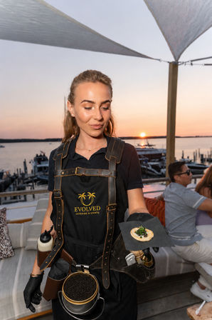 Server in a black apron presenting a caviar-topped canapé on a cracker at a sunset waterfront event by a marina with boats
