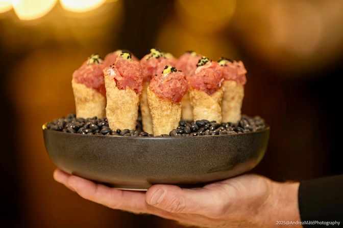 Close-up of gourmet tuna tartare cones — crispy pastry shells filled with raw tuna, topped with creamy aioli and black sesame, arranged in a bowl of black beans and held on a hand against a warm bokeh background.