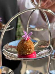 Golden breadcrumb-coated canapé on bright pink sauce, topped with a small purple edible flower, presented on a reflective silver tiered stand