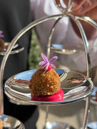 Golden breadcrumb-coated canapé on bright pink sauce, topped with a small purple edible flower, presented on a reflective silver tiered stand