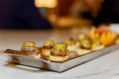 Close-up of gourmet mini burger sliders on round toasted bread topped with dill pickle slices, arranged on a metal serving tray on a marble countertop — bite-sized catering appetizers