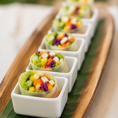 Row of bite-sized rainbow vegetable spring rolls with tofu, corn, carrots and purple cabbage served in white ceramic cups on a banana-leaf-lined wooden platter, fresh vegan appetizer