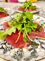 Vibrant tuna carpaccio: thin ruby slices topped with peppery arugula and microgreens, finished with herb vinaigrette on a decorative porcelain plate — gourmet restaurant appetizer.