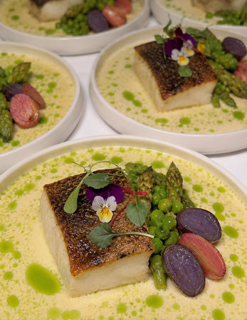 Seared white fish with crispy skin on a creamy herb sauce, plated with asparagus tips, green peas, purple and red baby potatoes, microgreens and edible viola flowers — vibrant gourmet restaurant entree