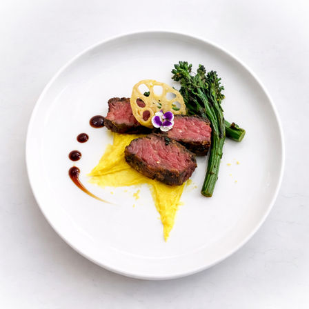 Plated medium-rare steak slices on a white plate with bright yellow vegetable puree, grilled broccolini, a lotus-root crisp, purple edible flower and balsamic sauce dots — minimalist fine-dining presentation