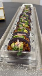 Row of bite-size blue corn tortilla tacos topped with guacamole, microgreens and a dollop of spicy crema, arranged on a clear acrylic serving tray — elegant catering appetizer.