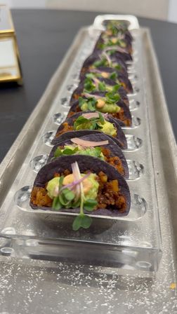 Row of bite-size blue corn tortilla tacos topped with guacamole, microgreens and a dollop of spicy crema, arranged on a clear acrylic serving tray — elegant catering appetizer.