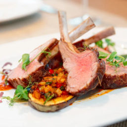 Juicy medium-rare rack of lamb chops with grilled eggplant, vibrant pepper relish and microgreens on a white plate — gourmet fine-dining dish.