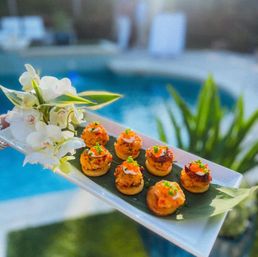 Poolside platter of eight bite-size savory canapés on a banana leaf, topped with bright orange garnish and microgreens, styled with white orchids — outdoor poolside appetizer shot.