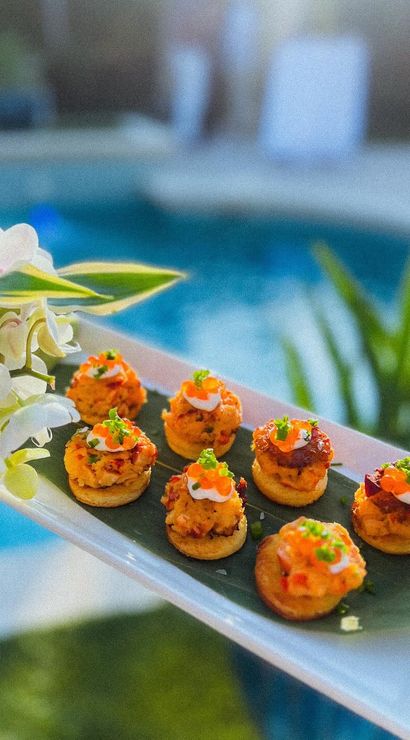 Poolside platter of eight bite-size savory canapés on a banana leaf, topped with bright orange garnish and microgreens, styled with white orchids — outdoor poolside appetizer shot.