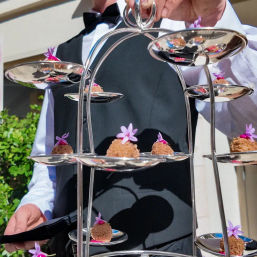 Server in black vest holding a silver tiered dessert stand of bite-sized chocolate truffles topped with small purple edible flowers, served outdoors at an event